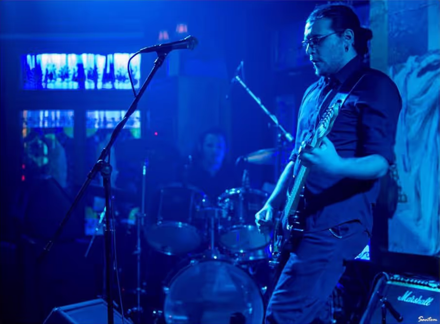 Petros passionately playing the guitar on stage with Alex on drums in the background, creating a dynamic live performance atmosphere.